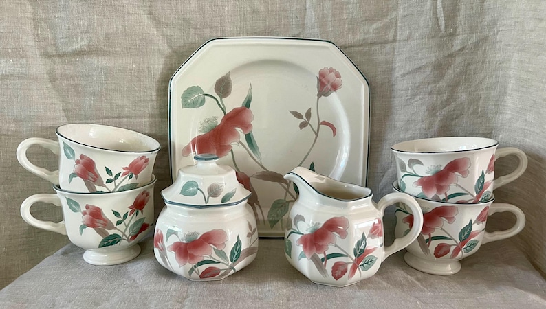 May include: A set of floral patterned teacups and saucers, a creamer, a sugar bowl, and a serving plate. The floral pattern features pink flowers and green leaves on a white background.