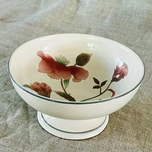 May include: A white pedestal bowl with a floral design. The bowl features a large pink flower with green leaves and a smaller pink flower on the side. The bowl has a green rim and a green band around the base.