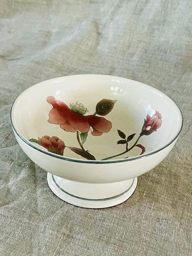 May include: A white pedestal bowl with a floral design. The bowl features a large pink flower with green leaves and a smaller pink flower on the side. The bowl has a green rim and a green band around the base.