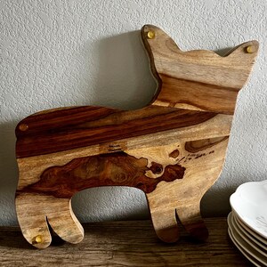 Op de afbeelding: Een houten snijplank in de vorm van een hond. De plank is gemaakt van licht en donkerbruin hout met een natuurlijk houtnerfpatroon. De plank heeft messing accenten op de oren en poten.