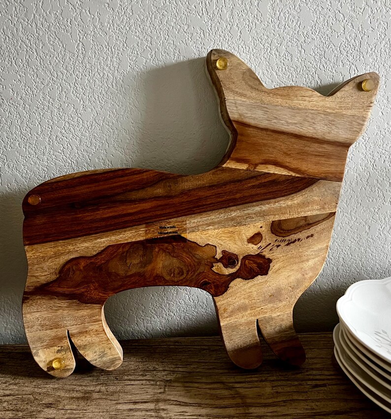 Op de afbeelding: Een houten snijplank in de vorm van een hond. De plank is gemaakt van licht en donkerbruin hout met een natuurlijk houtnerfpatroon. De plank heeft messing accenten op de oren en poten.