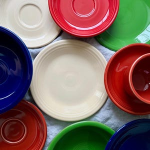 Puede incluir: Una colección de platos de cerámica de colores en rojo, verde, azul y blanco. Los platos están dispuestos sobre una superficie de tela gris claro.