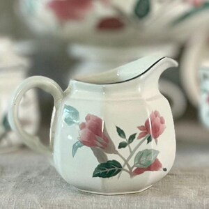 May include: White ceramic creamer with a floral design featuring pink roses and green leaves. The creamer has a rounded body and a curved handle.