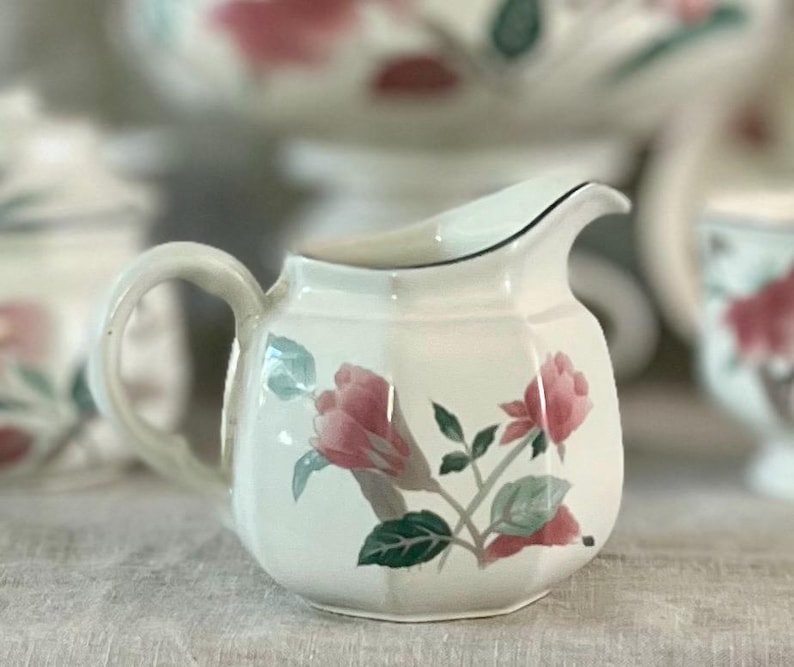 May include: White ceramic creamer with a floral design featuring pink roses and green leaves. The creamer has a rounded body and a curved handle.