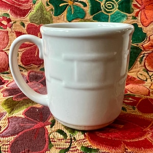 May include: White ceramic mug with a basket weave design. The mug has a large handle and is sitting on a colorful embroidered fabric.