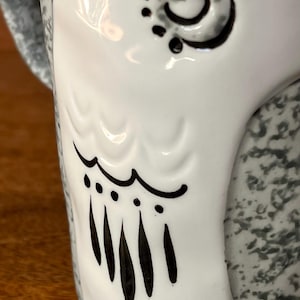 May include: Close-up of a white ceramic mug with a decorative owl design. The owl's eye and wing details are outlined in black. The mug's handle and the back of the mug have a speckled gray pattern.