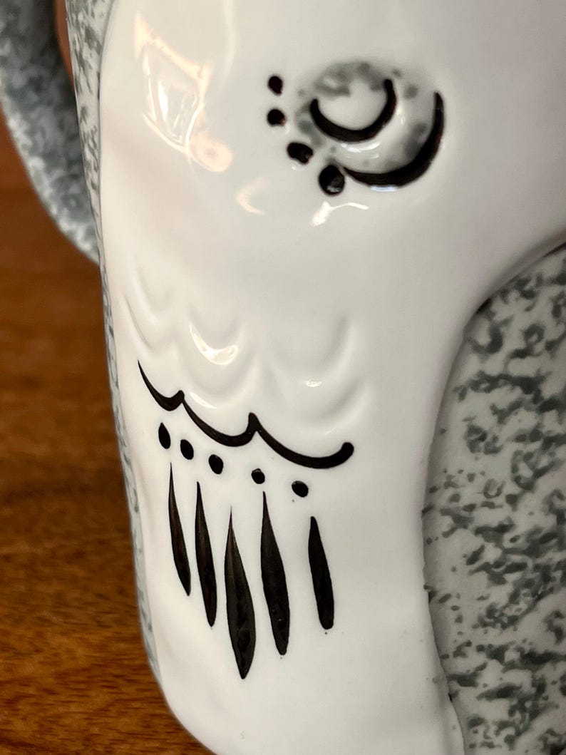 May include: Close-up of a white ceramic mug with a decorative owl design. The owl's eye and wing details are outlined in black. The mug's handle and the back of the mug have a speckled gray pattern.