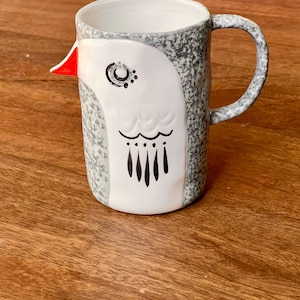 May include: A ceramic mug with a bird design. The mug features a white bird face with black and red accents against a speckled gray background. The handle is also gray and speckled. The mug is on a wooden surface.