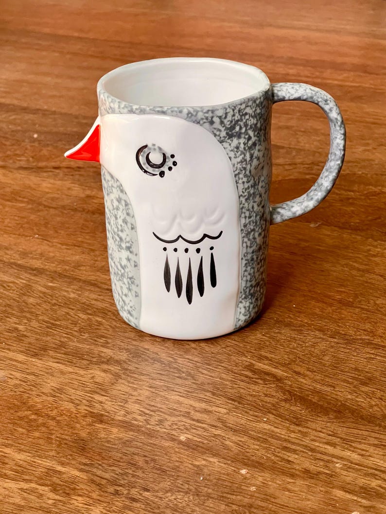 May include: A ceramic mug with a bird design. The mug features a white bird face with black and red accents against a speckled gray background. The handle is also gray and speckled. The mug is on a wooden surface.