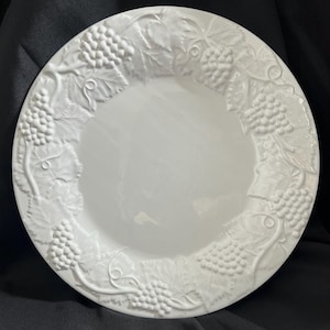 May include: A white ceramic plate with a raised grape and leaf design around the rim. The plate has a smooth, glossy surface and a slightly recessed center. The grape clusters and leaves are detailed and add a decorative touch to the plate.