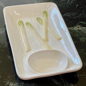May include: White ceramic serving dish with a small dipping bowl and four asparagus spears arranged on the plate.