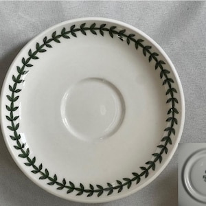 May include: White ceramic saucer with a green leafy border design. The saucer has a smaller, circular depression in the center.