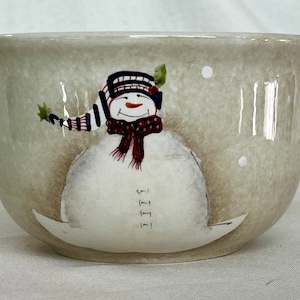 May include: A ceramic bowl with a snowman design. The snowman is wearing a red and white striped hat and a red scarf. The bowl has a beige background with white dots.