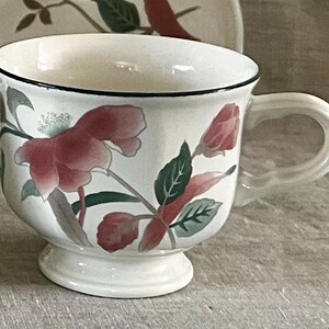 May include: A white teacup with a black rim and floral design. The flowers are pink, red, and green. The cup has a pedestal base.