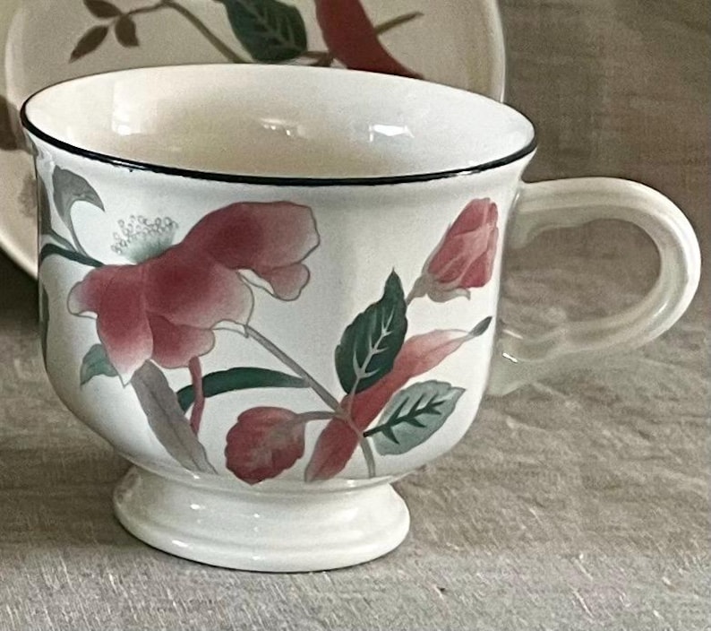 May include: A white teacup with a black rim and floral design. The flowers are pink, red, and green. The cup has a pedestal base.
