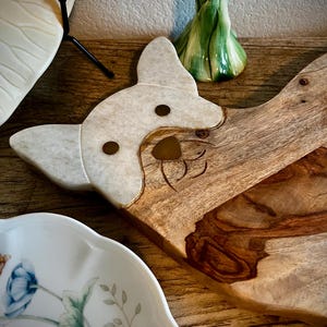 Op de afbeelding: Een houten snijplank met een wit marmeren hondenkopontwerp. De plank heeft een natuurlijke houtnerf en is perfect om kaas, crackers of andere hapjes op te serveren.