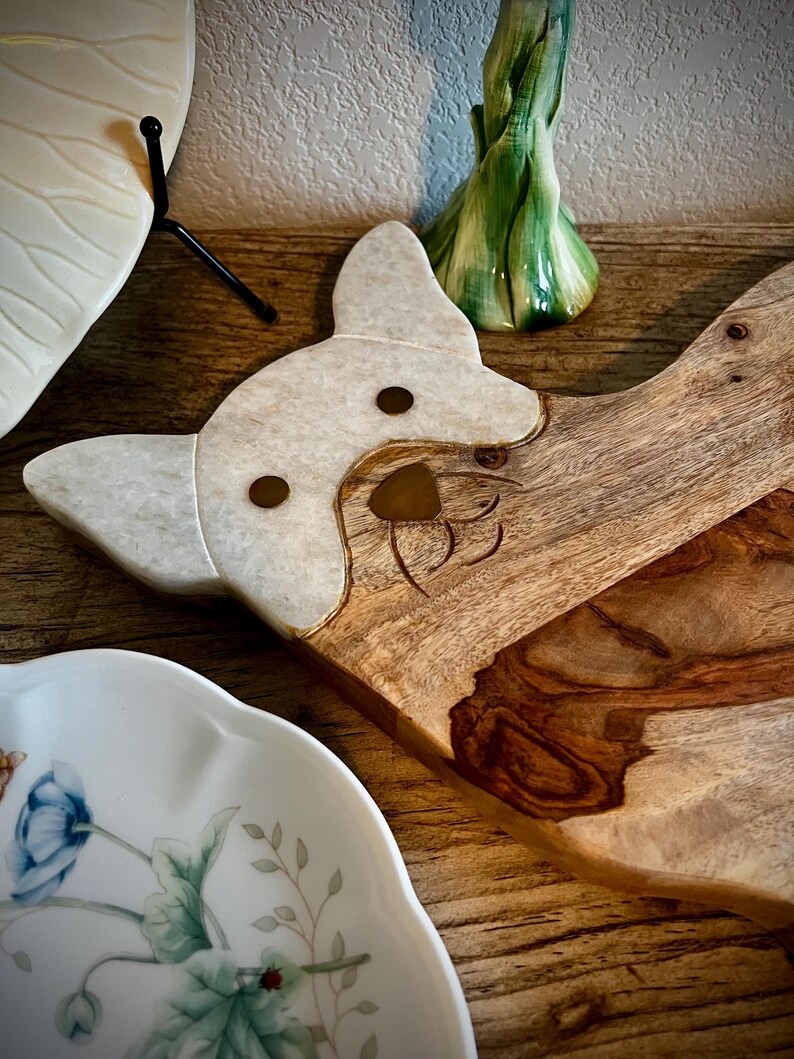 Op de afbeelding: Een houten snijplank met een wit marmeren hondenkopontwerp. De plank heeft een natuurlijke houtnerf en is perfect om kaas, crackers of andere hapjes op te serveren.