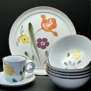 May include: A set of white ceramic dinnerware with a floral design featuring yellow, orange, purple, and green flowers. The set includes a large dinner plate, a smaller bowl, and a cup and saucer.