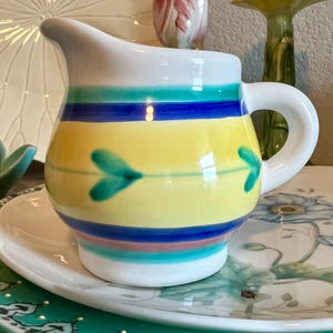 May include: A small, ceramic pitcher with a white handle and spout. The pitcher is decorated with horizontal bands of turquoise, blue, and pink over a yellow base. Two teal arrow-shaped designs are painted on the yellow section. The pitcher sits on a white saucer.