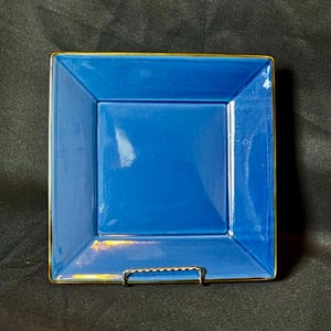 May include: A square, cobalt blue plate with a gold trim. The plate is made of ceramic and has a glossy finish. The plate is displayed on a metal stand against a dark background.