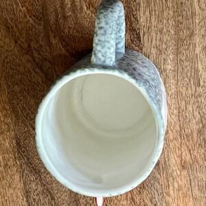 May include: A speckled ceramic mug with a curved handle. The mug's exterior features a gray and white pattern, while the interior is a light cream color. The mug is set against a wooden surface.
