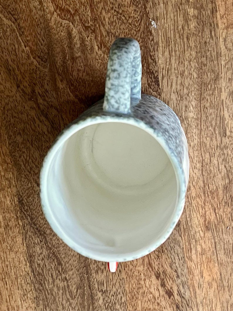 May include: A speckled ceramic mug with a curved handle. The mug's exterior features a gray and white pattern, while the interior is a light cream color. The mug is set against a wooden surface.