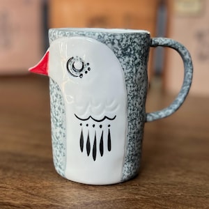 May include: A ceramic mug with a bird design. The mug features a white bird face with a red beak, black eyes, and a black pattern. The body of the mug is speckled gray and white, with a matching handle. The mug is on a wooden surface.