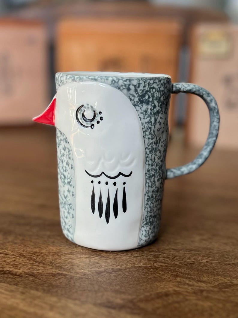 May include: A ceramic mug with a bird design. The mug features a white bird face with a red beak, black eyes, and a black pattern. The body of the mug is speckled gray and white, with a matching handle. The mug is on a wooden surface.