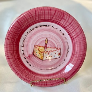 May include: Pink ceramic plate with a hand-painted design of a birthday cake with a single lit candle. The plate has a textured, woven pattern.