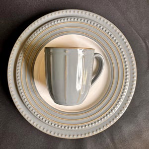 May include: A light gray ceramic mug with vertical ridges sits centered on a matching round plate. The plate features a raised beaded rim and a thin gold-toned circular accent line. Both pieces have a smooth, glossy finish and a neutral color palette.