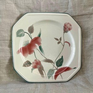 May include: A white square dinner plate with a green rim features a floral design of red and green flowers and leaves.