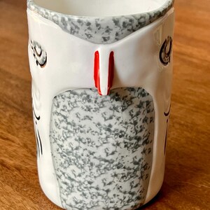 May include: A ceramic mug with a white exterior, featuring a gray speckled design and a red accent. The mug has a unique shape and a decorative pattern, perfect for coffee or tea.