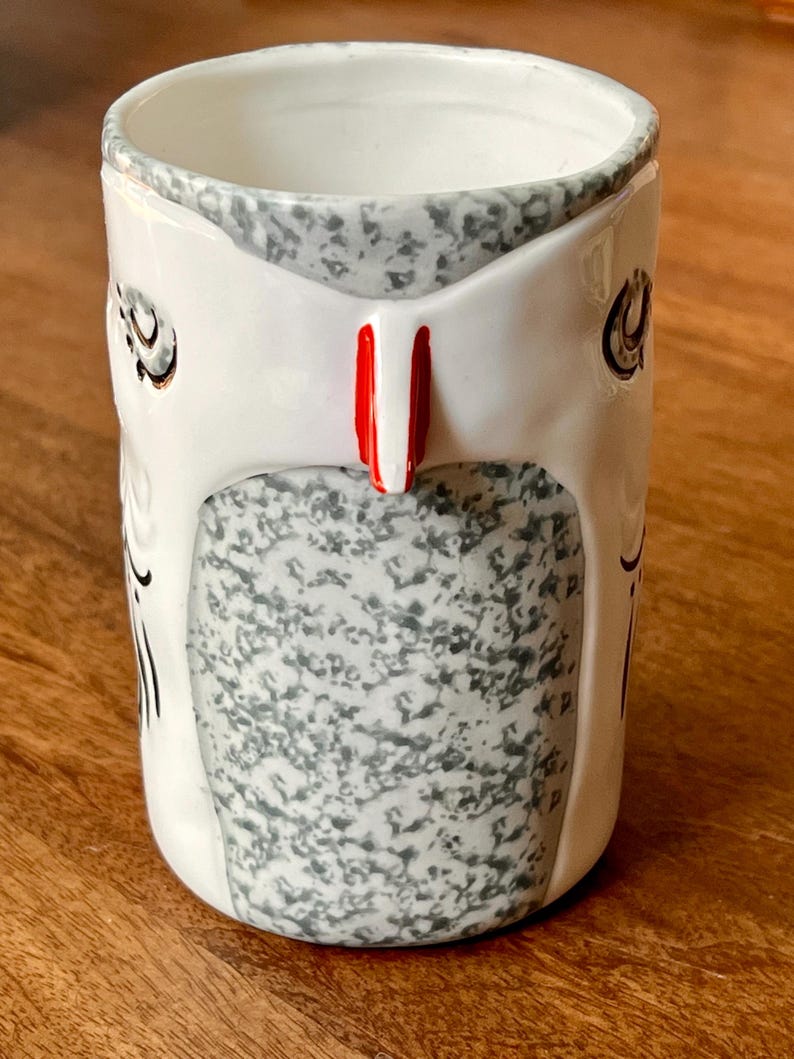May include: A ceramic mug with a white exterior, featuring a gray speckled design and a red accent. The mug has a unique shape and a decorative pattern, perfect for coffee or tea.