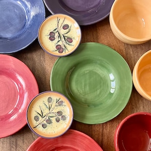 May include: A collection of colorful ceramic dishes and bowls. The set includes plates in shades of blue, purple, green, and red, along with smaller olive branch-decorated plates and orange and red bowls. The arrangement is on a wooden surface.