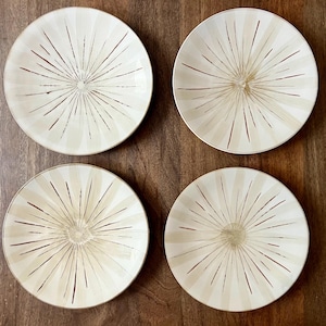 May include: Four off-white ceramic plates with a sunburst design in brown. The plates are round with a slightly raised edge and are arranged on a dark wood surface. The plates are a set for dining.