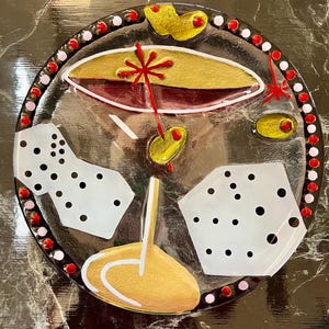 May include: A round glass plate with a gold and red design. The plate features a stylized martini glass with a red drink and a gold stem. The plate is decorated with white polka dots and a gold rim.