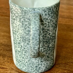 May include: A ceramic mug with a speckled, gray-green exterior and a white interior. The mug has a rounded handle and a slightly flared rim. The mug is sitting on a wooden surface.