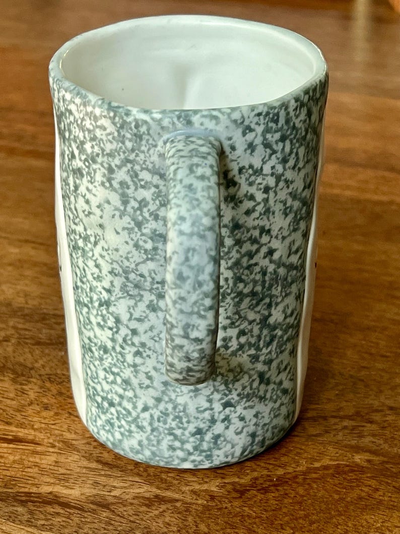 May include: A ceramic mug with a speckled, gray-green exterior and a white interior. The mug has a rounded handle and a slightly flared rim. The mug is sitting on a wooden surface.