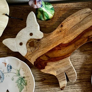 Op de afbeelding: Een houten snijplank in de vorm van een hond met een wit gezicht en bruine oren. De plank is gemaakt van natuurlijk hout met een lichtbruine kleur en een donkerbruine nerf.