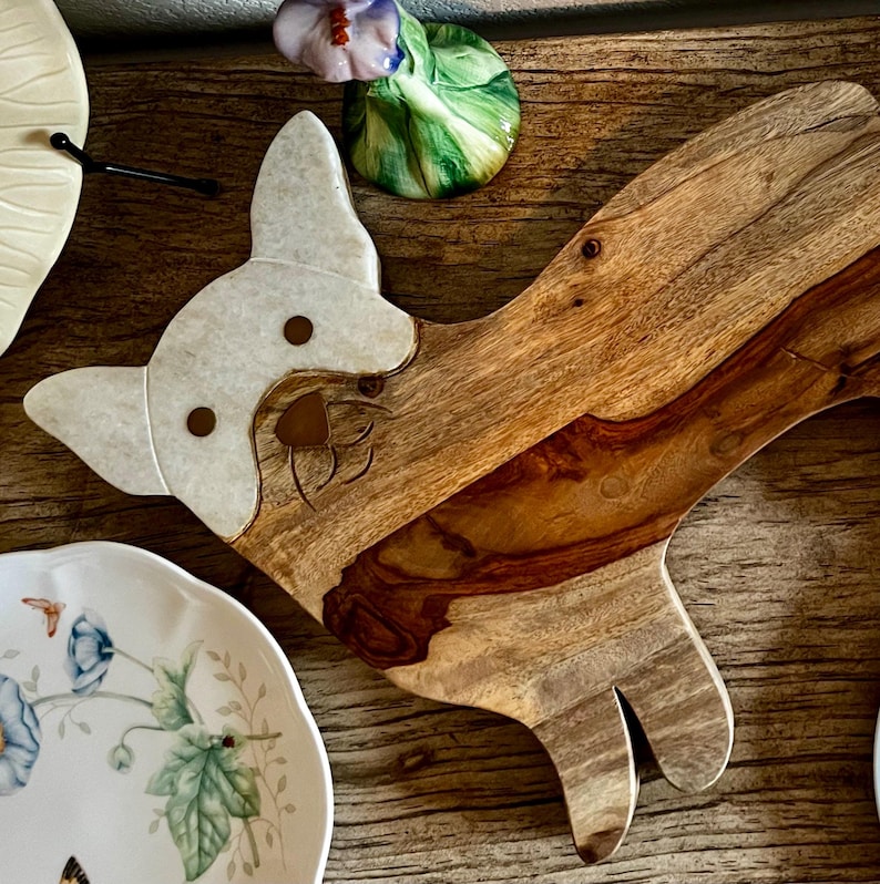 Op de afbeelding: Een houten snijplank in de vorm van een hond met een wit gezicht en bruine oren. De plank is gemaakt van natuurlijk hout met een lichtbruine kleur en een donkerbruine nerf.
