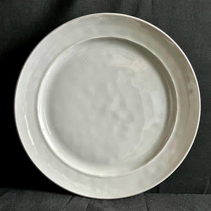 May include: A round, light gray ceramic plate with a wide rim. The plate has a slightly textured surface and a simple, elegant design. Ideal for serving meals or as a decorative piece. The plate is approximately 10 inches in diameter.