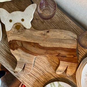 Op de afbeelding: Een houten snijplank in de vorm van een hond. De plank is gemaakt van licht en donker hout en heeft een witte en gouden inleg in de vorm van een hondenkop. De plank ligt op een houten tafel.