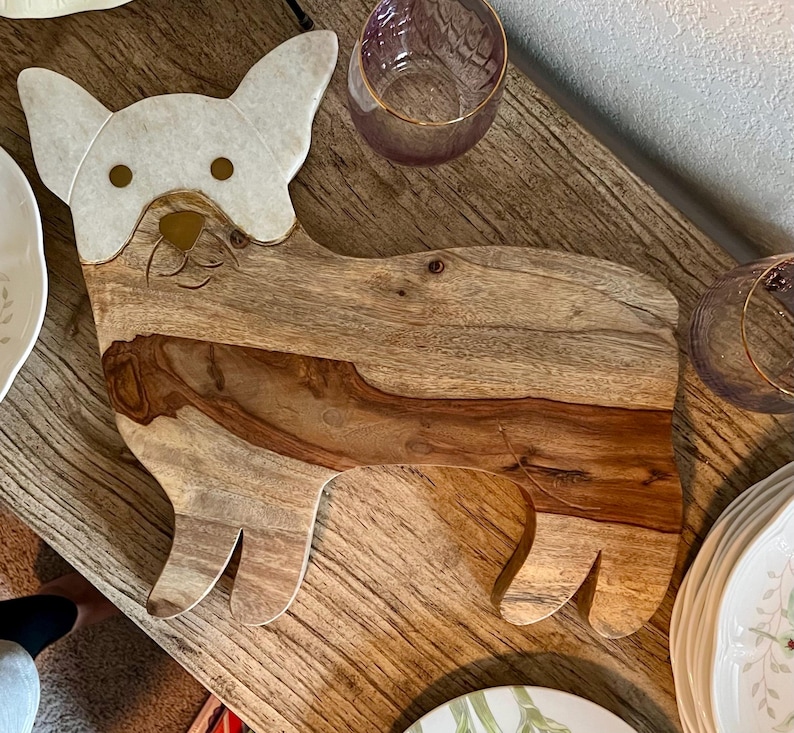 Op de afbeelding: Een houten snijplank in de vorm van een hond. De plank is gemaakt van licht en donker hout en heeft een witte en gouden inleg in de vorm van een hondenkop. De plank ligt op een houten tafel.