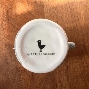 May include: White ceramic mug base with a black bird graphic and the text "by ANTHROPOLOGIE". The mug has a gray and white speckled design around the rim and handle. The mug is sitting on a wooden surface.