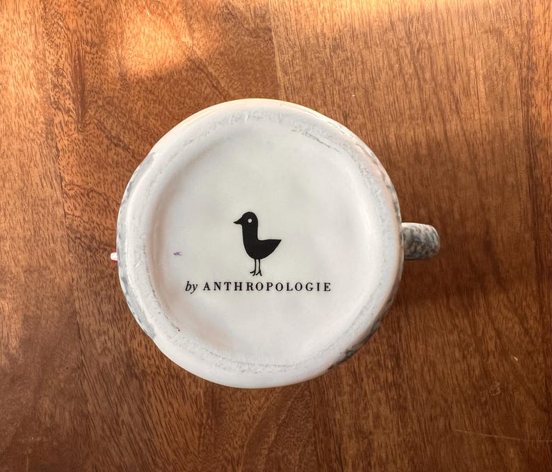 May include: White ceramic mug base with a black bird graphic and the text "by ANTHROPOLOGIE". The mug has a gray and white speckled design around the rim and handle. The mug is sitting on a wooden surface.