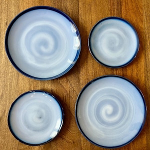 May include: Four ceramic plates with a blue and white swirl design. The plates have a dark blue rim and are arranged on a wooden surface. The plates vary in size, with two larger and two smaller plates.
