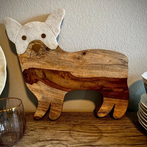 Op de afbeelding: Een houten snijplank in de vorm van een hond, met een wit geschilderd gezicht en oren. De plank heeft een natuurlijke houtnerf en is perfect om kaas of charcuterie op te serveren.