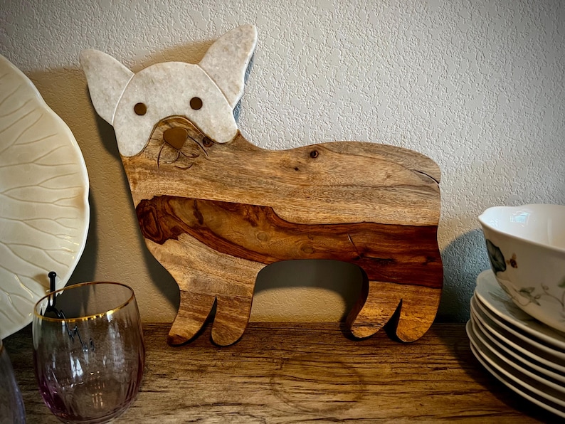 Op de afbeelding: Een houten snijplank in de vorm van een hond, met een wit geschilderd gezicht en oren. De plank heeft een natuurlijke houtnerf en is perfect om kaas of charcuterie op te serveren.