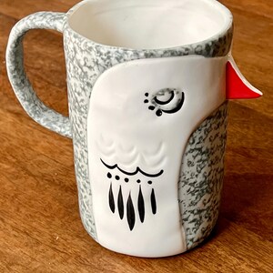 May include: A ceramic mug with a bird design. The mug features a white bird face with black details and a red beak. The body of the mug is speckled gray and white, with a matching handle.