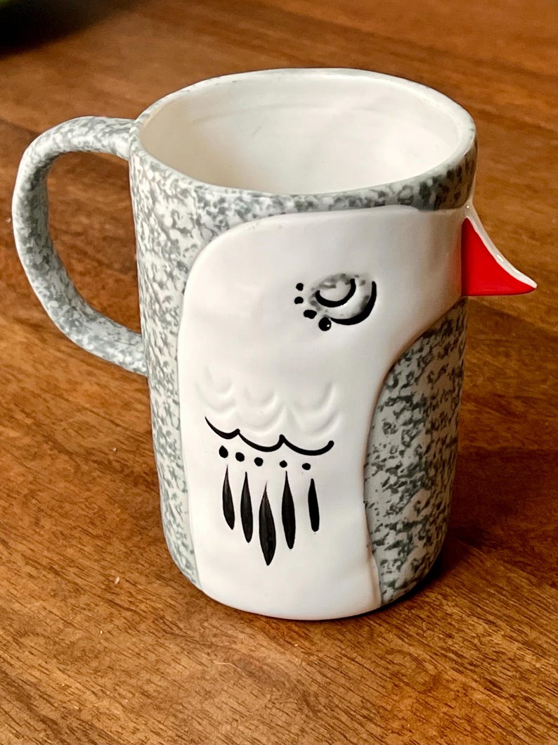 May include: A ceramic mug with a bird design. The mug features a white bird face with black details and a red beak. The body of the mug is speckled gray and white, with a matching handle.